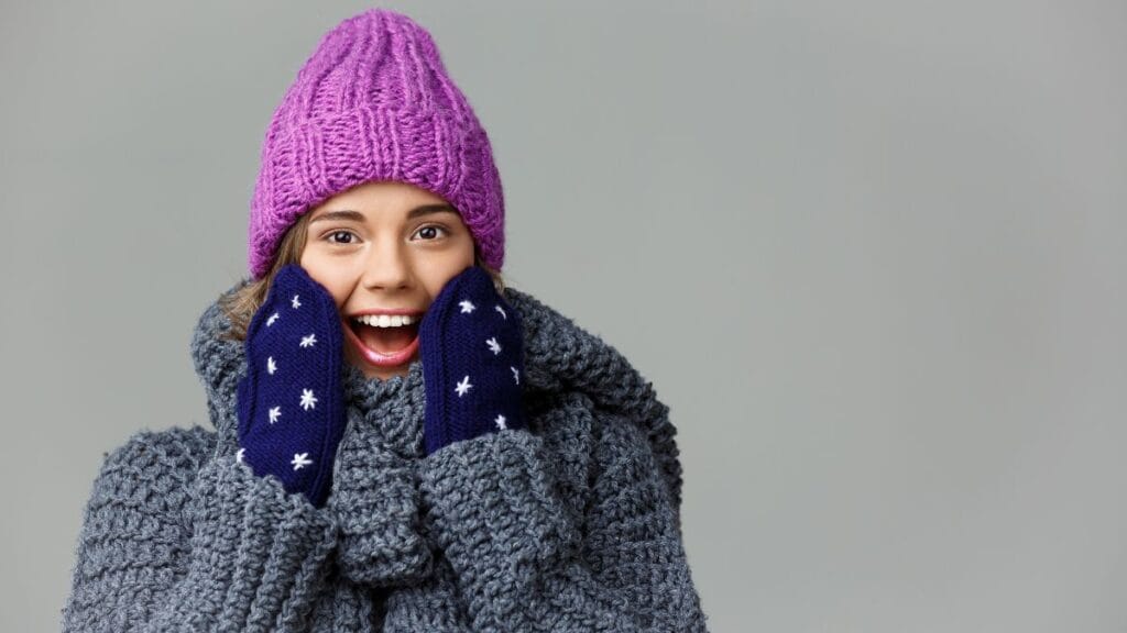 A young woman smiles widely while wearing a purple knitted hat, navy gloves with white star patterns, and a thick gray scarf. She appears excited, holding her hands to her face in a cheerful pose.