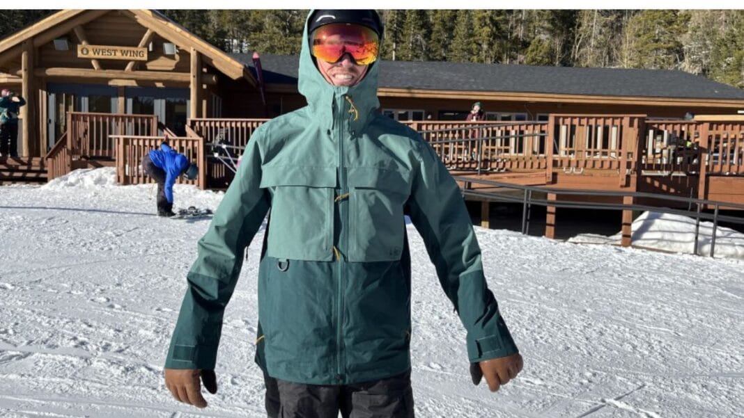 Man wearing a teal men's ski jacket with goggles, standing on snow with a ski resort cabin in the background.