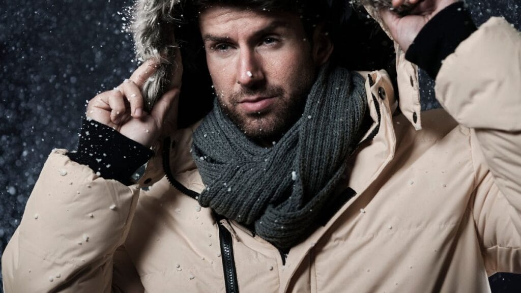 A man in a beige winter jacket and grey scarf, adjusting his fur-lined hood in the snow.