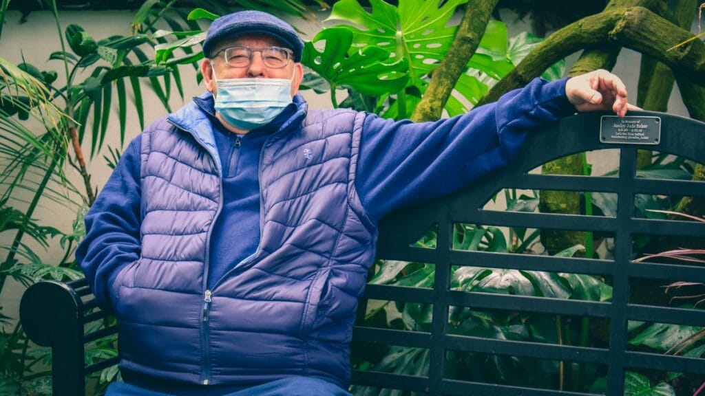 An elderly man wearing a blue puffer jacket, cap, and a mask, sitting on a black park bench surrounded by plants. There is a plaque on the bench in memory of someone.