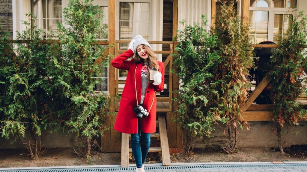 A smiling woman wearing a red coat, gray sweater, white hat, and gloves, holding a coffee cup and a camera, standing outdoors in front of a wooden house with green shrubs.