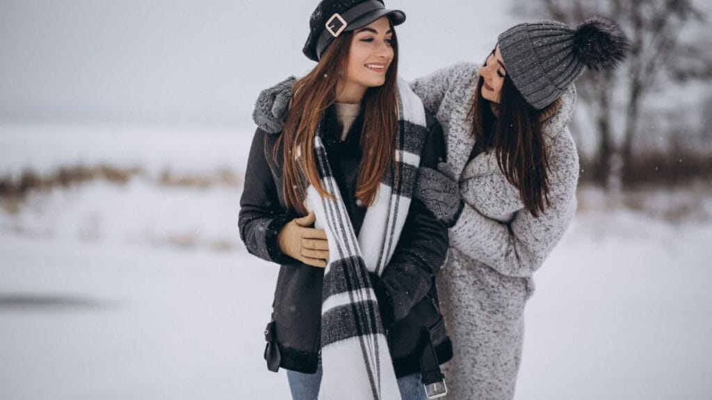 Two women smiling and standing together in a snowy landscape, both wearing stylish cold weather clothes. One woman is dressed in a black jacket with a scarf and a black hat, while the other wears a gray sweater, gray hat with a pom-pom, and a scarf.