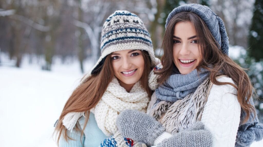 Two young women outdoors in a snowy setting, smiling while wearing winter hats, scarves, and warm layered clothing.