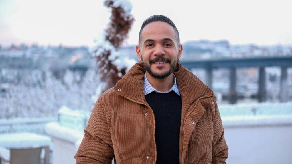 A man stands outdoors, smiling in a cozy brown corduroy winter jacket with a snow-covered landscape in the background.