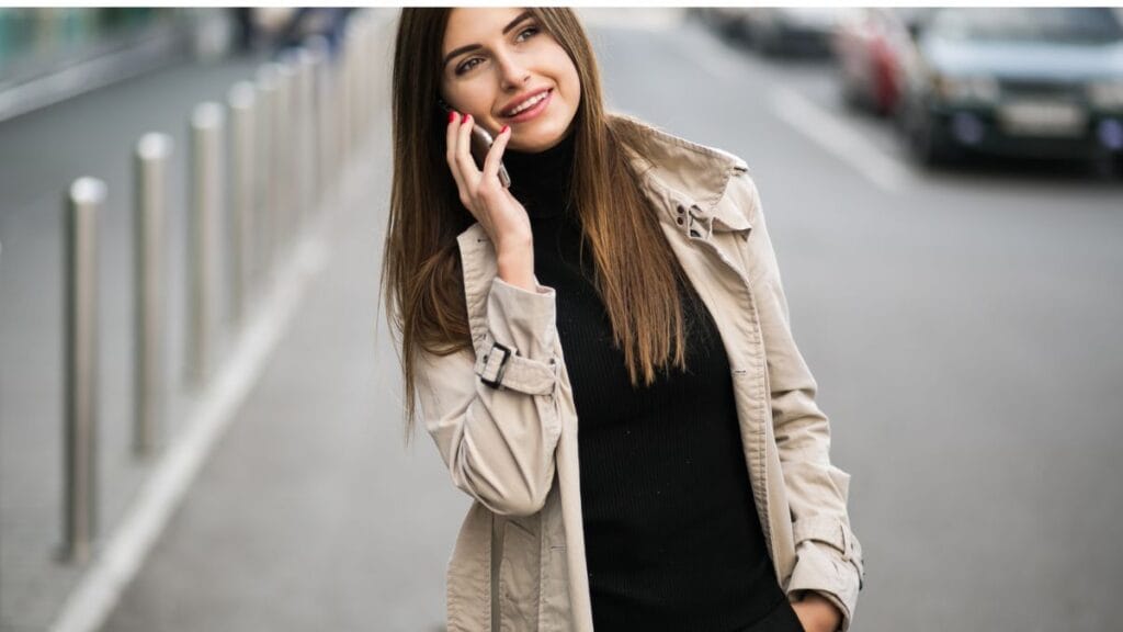 A woman wearing a beige jacket and black turtleneck, smiling while talking on the phone outdoors.