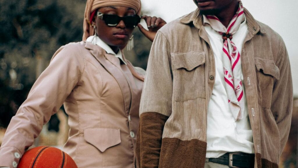 A fashionable woman and man posing together. The woman is holding a basketball and wearing a beige jacket with a headscarf, while the man is in a corduroy jacket and scarf.