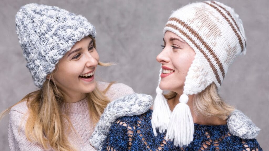 Two young women are smiling at each other, wearing cozy knitted hats and gloves. One woman is wearing a gray and white hat, while the other is wearing a white hat with brown stripes and tassels. They are both dressed warmly in sweaters, standing against a neutral gray background.