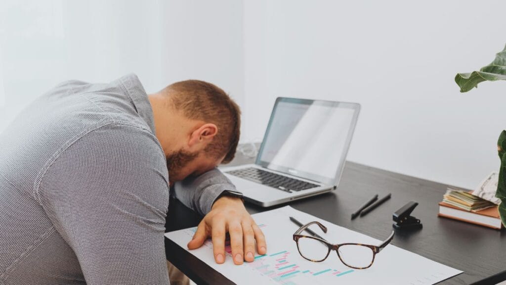 A person sitting at a desk, feeling overwhelmed, with their head resting on the table next to a laptop and papers.