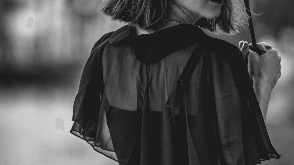 Woman in a black sheer cape standing with her back to the camera, holding an umbrella, with a blurred background.