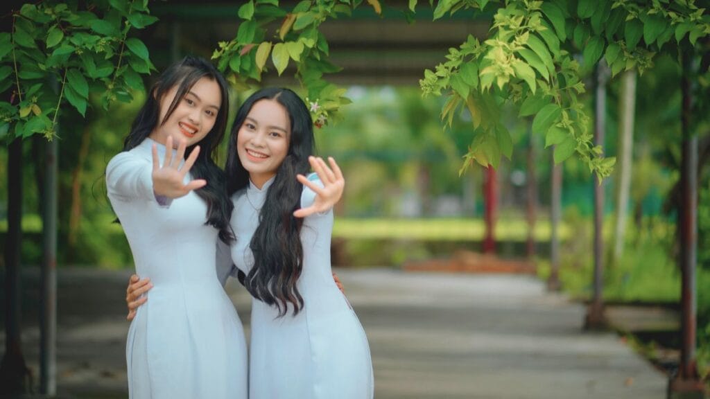 Two women in traditional white dresses smile and pose playfully, surrounded by green foliage.