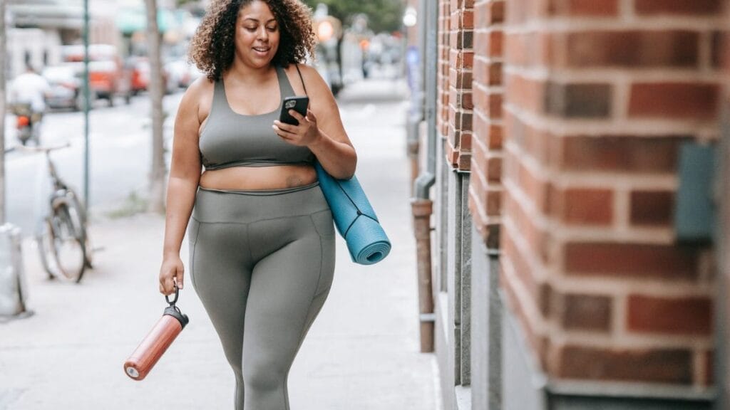 The image shows a woman wearing a matching olive green sports bra and leggings, walking outside on a city sidewalk. She is holding a water bottle in one hand and a yoga mat rolled up in the other. She is looking at her phone, smiling, with curly hair and a relaxed, active look. The background features a brick wall and some trees.