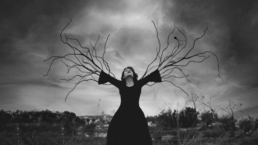 Woman in a black dress with surreal, branch-like extensions emerging from her arms, set against a dramatic cloudy sky.