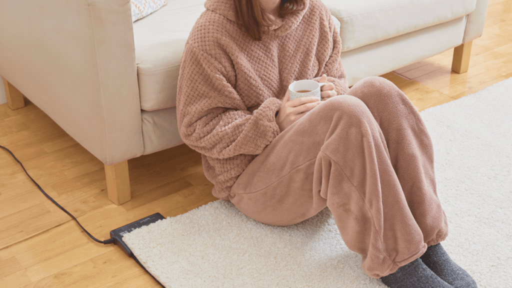 A person sitting on the floor in cozy brown loungewear, holding a cup, with a soft rug and a heated mat nearby.