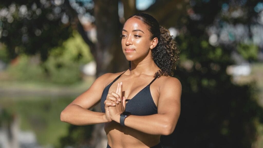 Woman wearing a black sports bra, practicing yoga outdoors, with her hands in a prayer position and a serene expression.