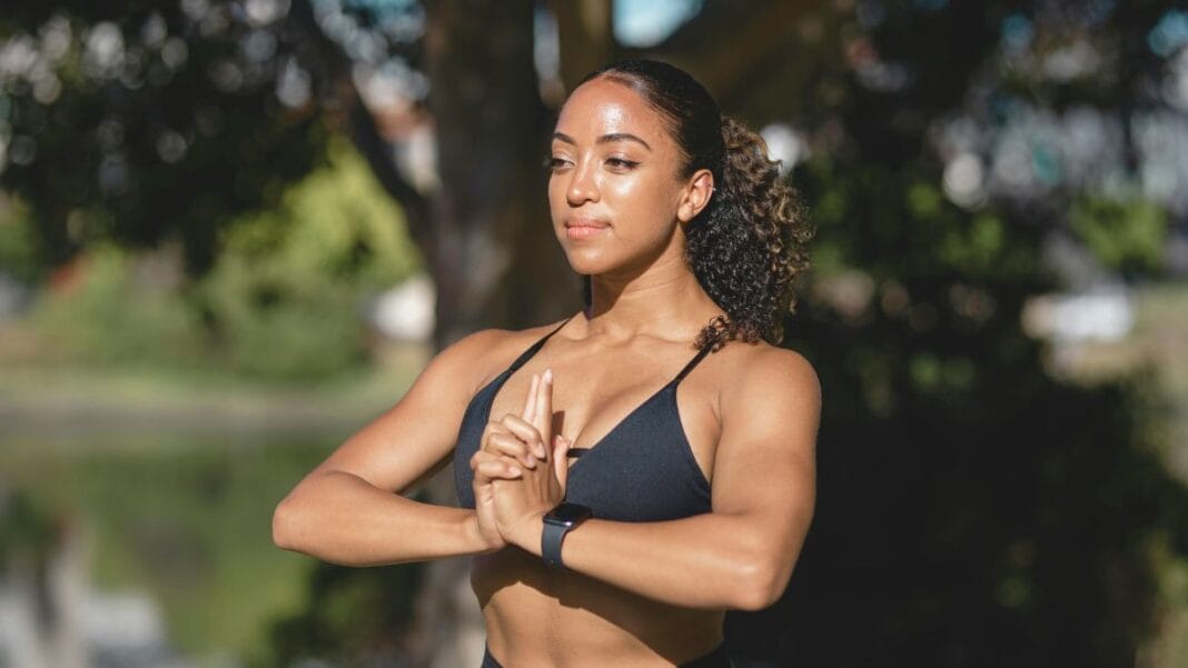 Woman wearing a black sports bra, practicing yoga outdoors, with her hands in a prayer position and a serene expression.