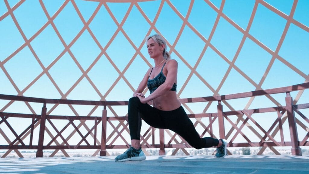 Woman in activewear performing a lunging stretch under a geometric structure, with a blue sky in the background.