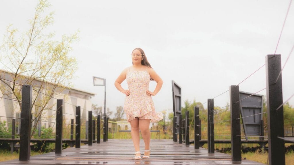 A confident woman stands on a wooden bridge, wearing a floral dress and sandals, with trees and buildings in the background.
