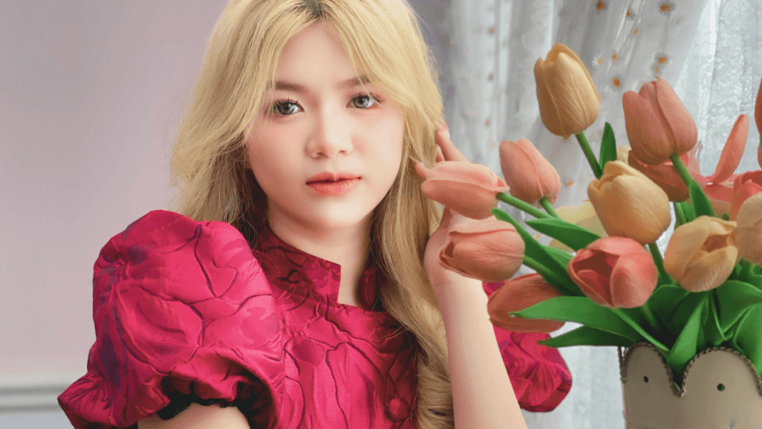 Young woman with blonde hair wearing a pink floral dress, posing next to a bouquet of peach-colored tulips with a soft expression