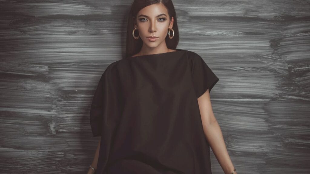 Woman in a black oversized dress, with large hoop earrings, posing against a textured dark grey background.
