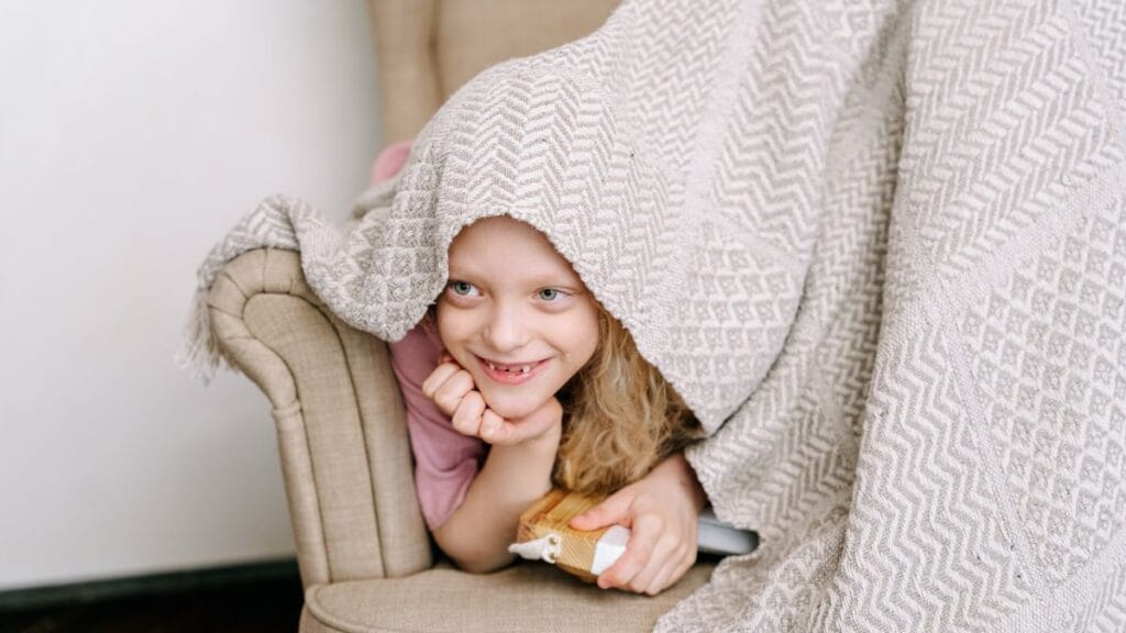 A young girl playfully peeking out from under a cozy beige blanket while sitting on a chair, smiling and resting her chin on her hand.