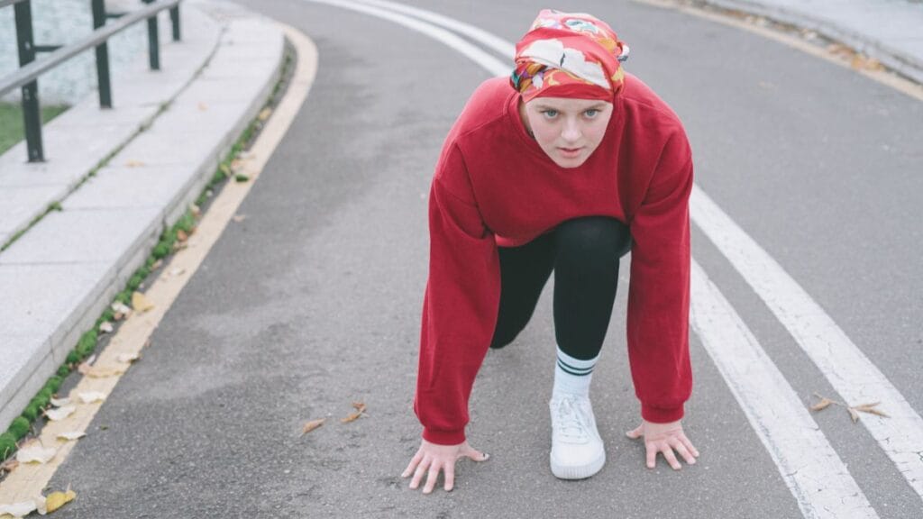 A woman in a red sweatshirt and leggings, crouching at the starting line on a road, preparing to run.