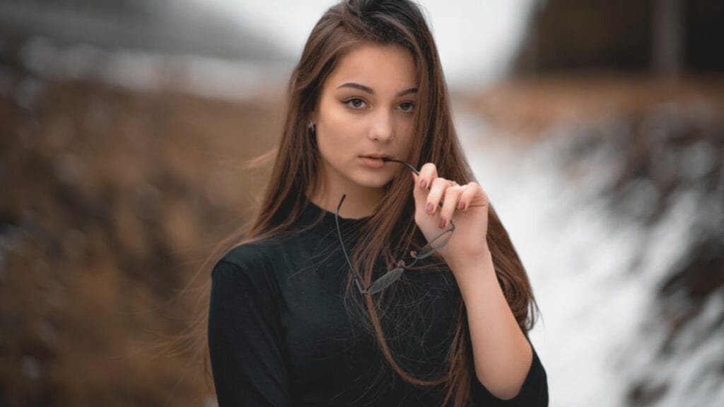 Young woman with long brown hair, wearing a black shirt, holding sunglasses in her mouth, looking at the camera with a serious expression.