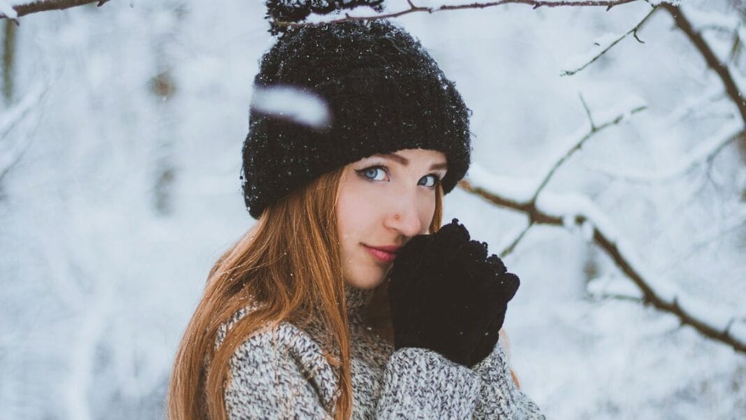 A woman in a black knitted hat and gloves, standing in the snow with her hands near her face, gazing into the camera.