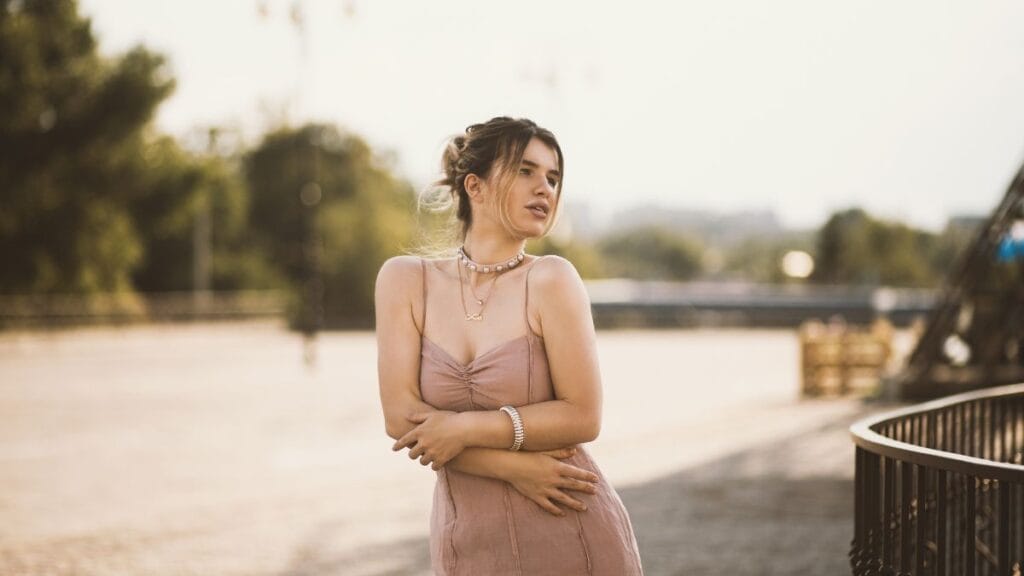  A woman in a blush pink dress stands outdoors, wearing a necklace and bracelet, gazing thoughtfully.