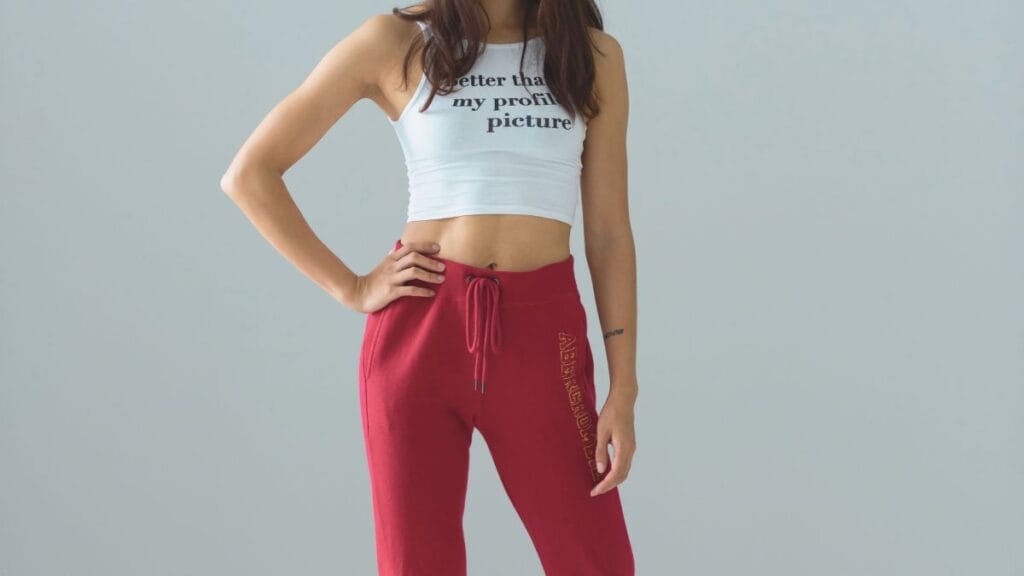 A woman wearing a white cropped top with the text "Better than my profile picture" and red sweatpants with a drawstring.