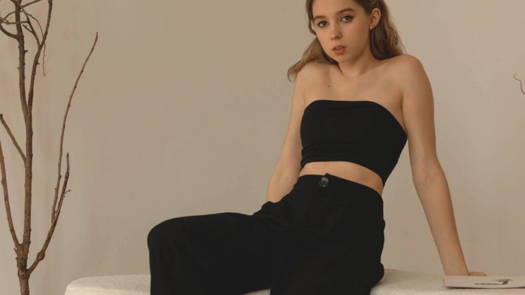 A woman wearing a black strapless top and black pants, sitting on a white surface with a minimalist background and a tree branch beside her.