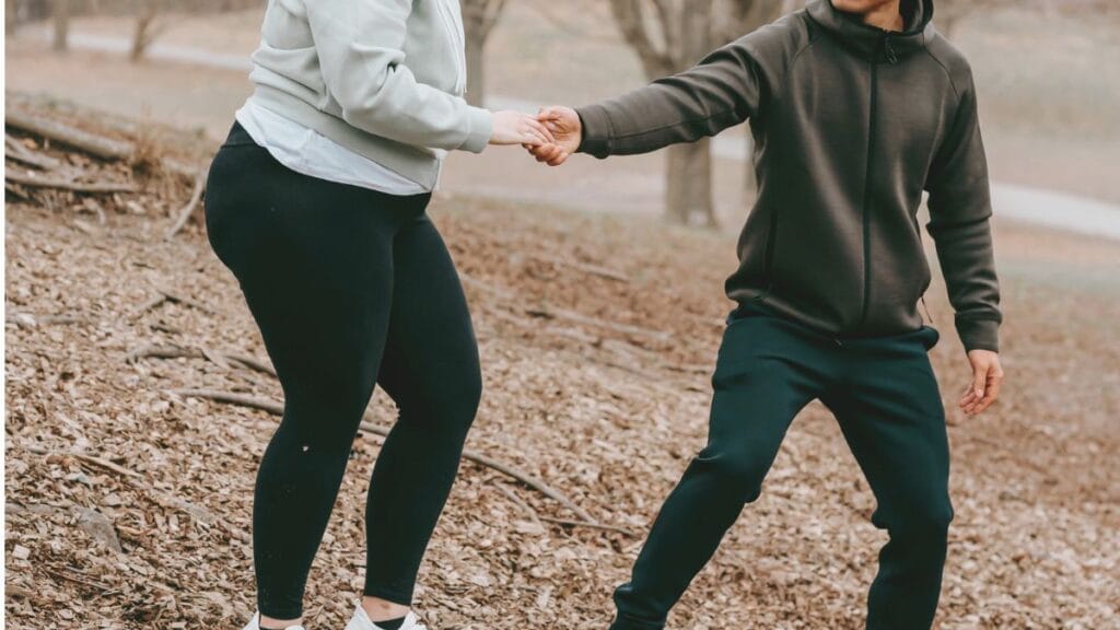 A plus-size woman wearing black leggings and a light gray hoodie holding hands with a man in a dark hoodie while walking outdoors.
