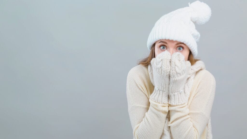 Woman wearing white winter clothes, hat, and gloves, covering her face with her hands in a cold-weather pose.