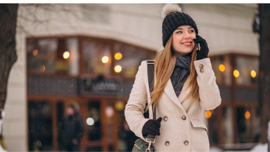 A woman wearing a winter going outfit with a beige coat, black knit beanie, scarf, and gloves while talking on her phone outdoors.