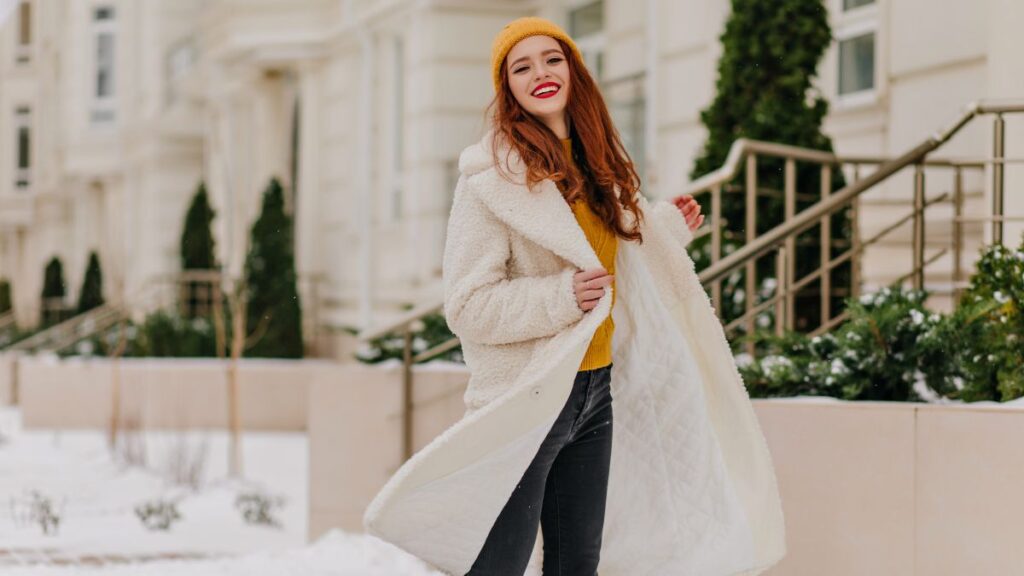 A young woman with red hair, wearing a white fluffy coat and yellow beanie, smiling while walking in the snow.
