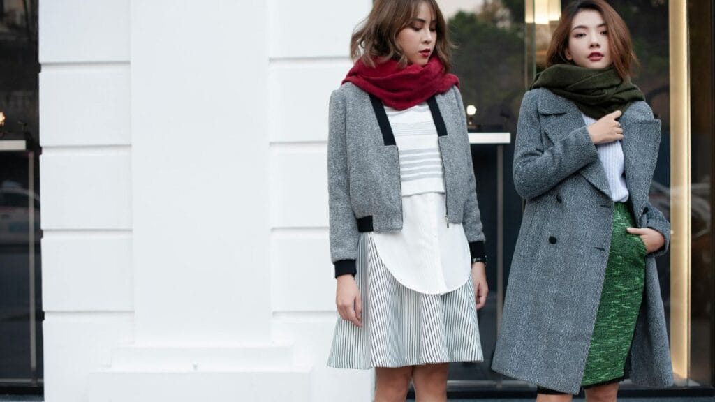 Two women dressed in stylish winter outfits, featuring scarves and coats, standing in front of a white building.