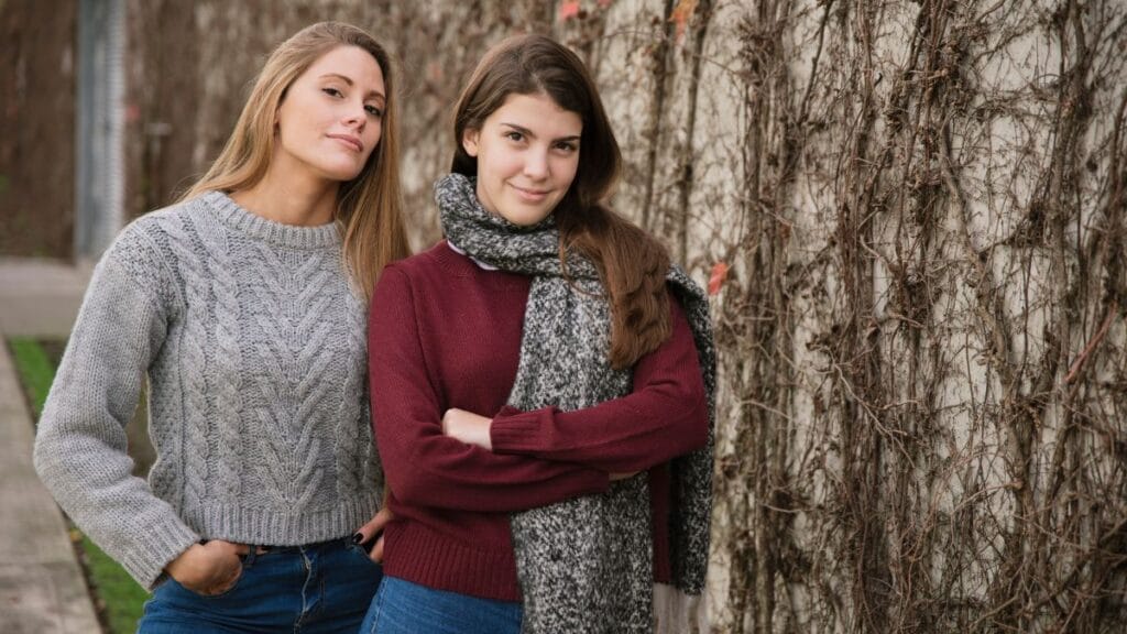 Two women standing side by side outdoors, wearing cozy winter sweaters with one woman in a gray cable-knit sweater and the other in a maroon sweater with a gray scarf, with a vine-covered wall in the background.