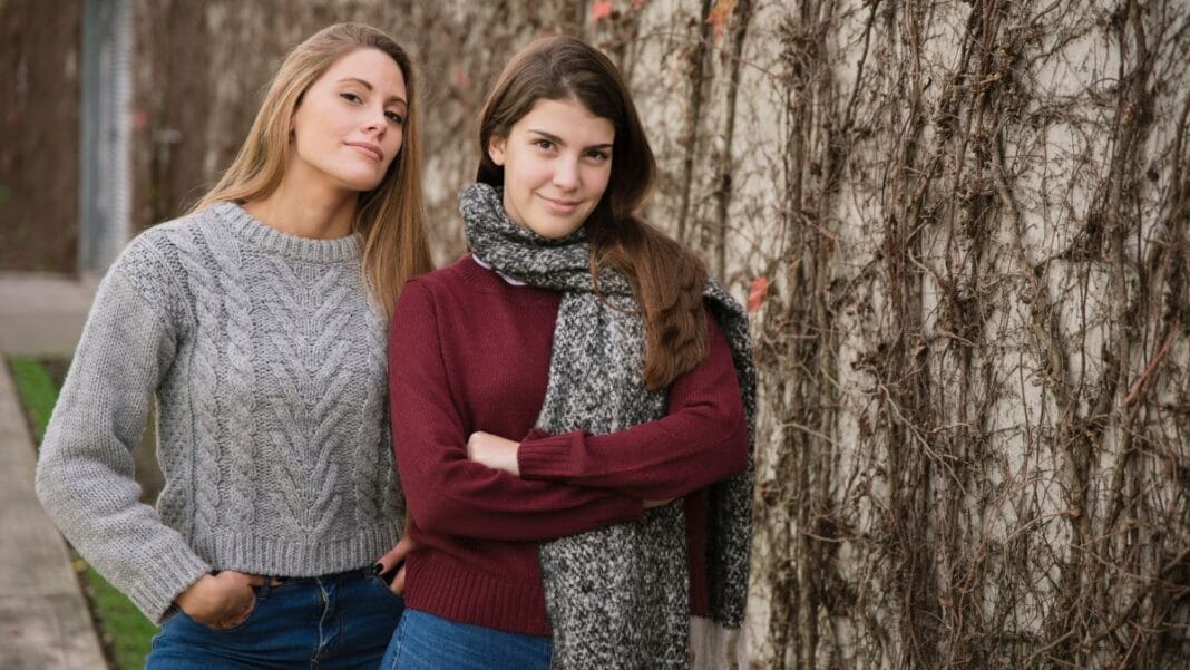 Two women standing side by side outdoors, wearing cozy winter sweaters with one woman in a gray cable-knit sweater and the other in a maroon sweater with a gray scarf, with a vine-covered wall in the background.