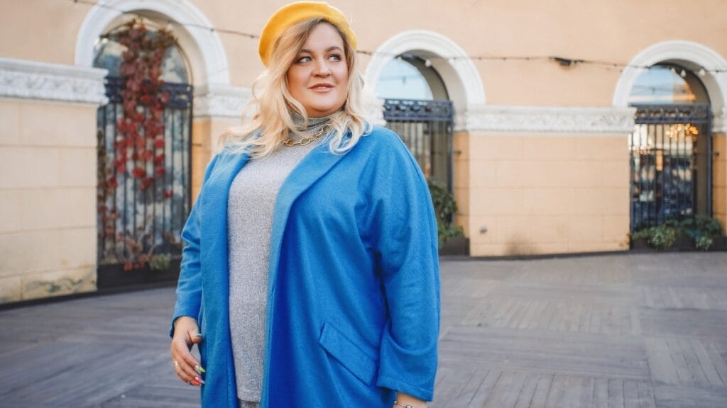 Plus size woman in a blue coat and yellow beret standing outside in front of an architectural backdrop