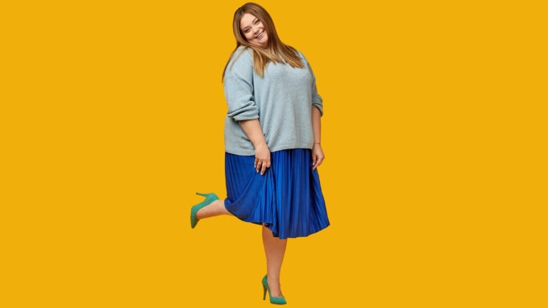 Plus size woman wearing a light blue sweater and blue pleated skirt, smiling with a bright yellow background