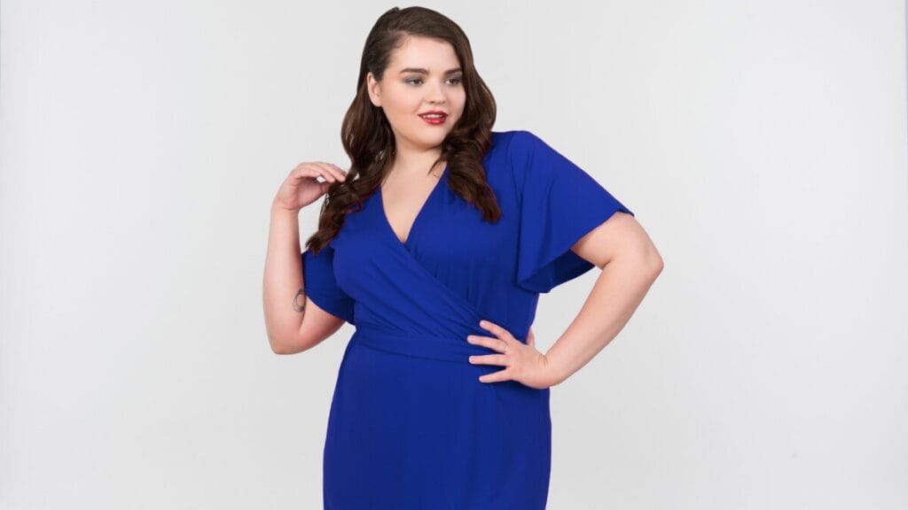 Plus-size woman wearing a blue wrap dress, posing confidently with one hand on her hip.