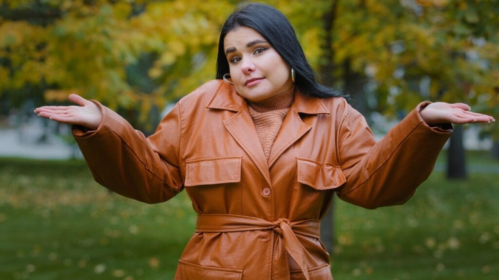 A woman in a brown plus size dress standing with her arms raised in a shrug, set against a fall backdrop
