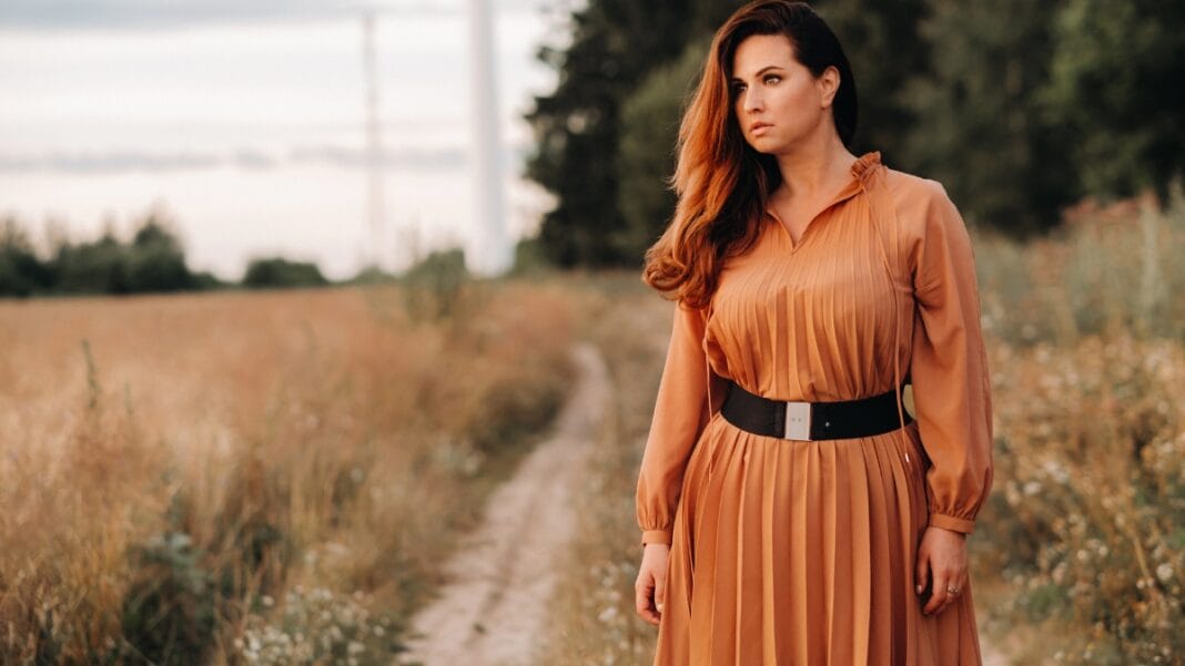 A woman in a flowing brown plus size dress standing in a field with a serene expression, showcasing the dress's flattering fit