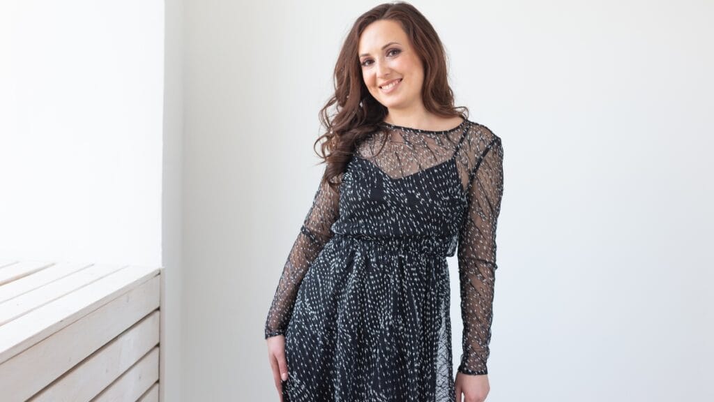 Curvy woman in a black sheer long-sleeve cocktail dress, size 14, standing near a window with natural light