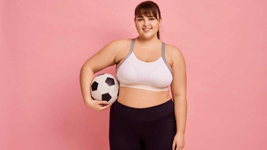 Woman wearing a white full figure sports bra, holding a soccer ball in front of a pink background