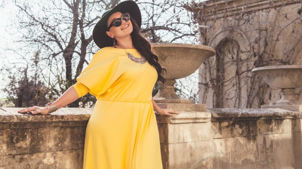 A woman in a yellow dress posing outdoors near a stone structure, smiling with a hat and stylish accessories