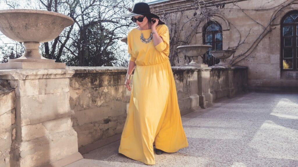 A woman in a yellow dress walking near a stone wall with accessories, showcasing a fashionable plus-size evening outfit