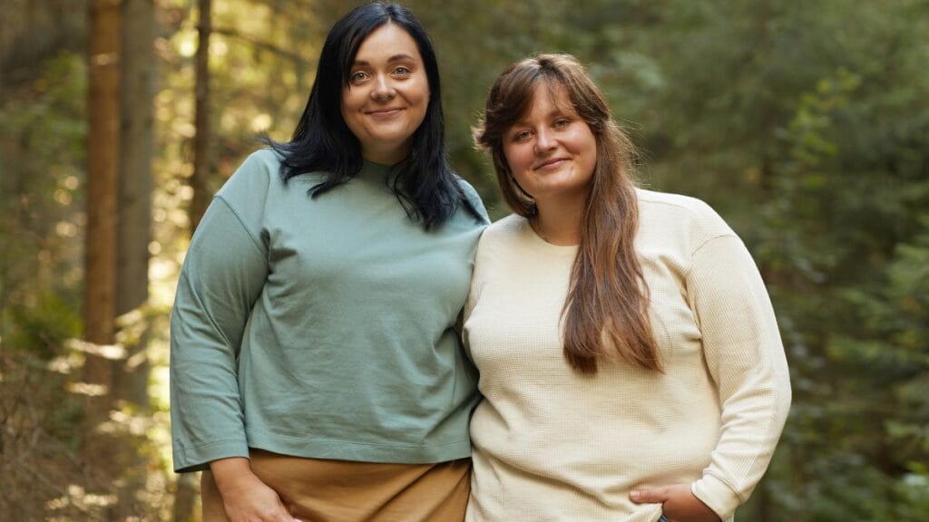 Two women posing outdoors in plus size sweaters, one in green and the other in cream