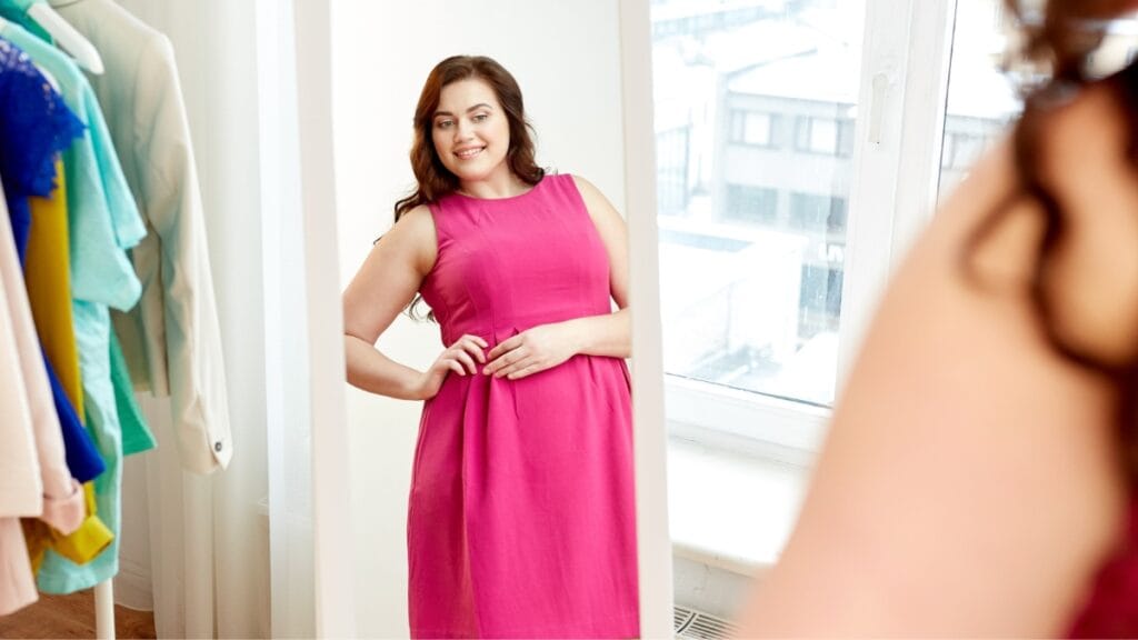 Plus size woman trying on a pink evening dress, looking at her reflection in a mirror