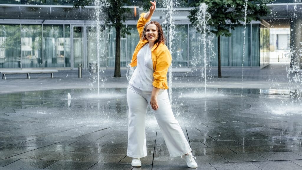 Plus size woman in a white tea dress with a yellow jacket and sneakers standing by a fountain