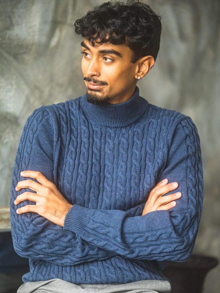 Man wearing a blue cable-knit turtleneck sweater with arms crossed, looking to the side in a relaxed indoor setting.
